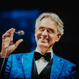 Andrea Bocelli w niebieskiej marynarce z czarną muszką, trzymający mikrofon.