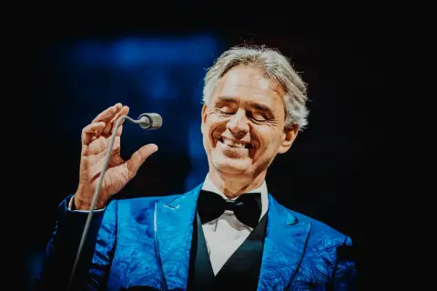 Andrea Bocelli w niebieskiej marynarce z czarną muszką, trzymający mikrofon.