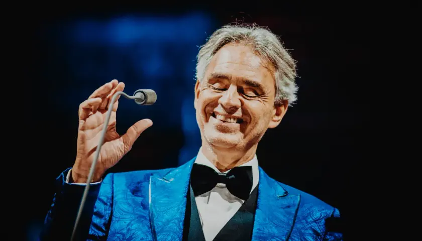 Andrea Bocelli w niebieskiej marynarce z czarną muszką, trzymający mikrofon.